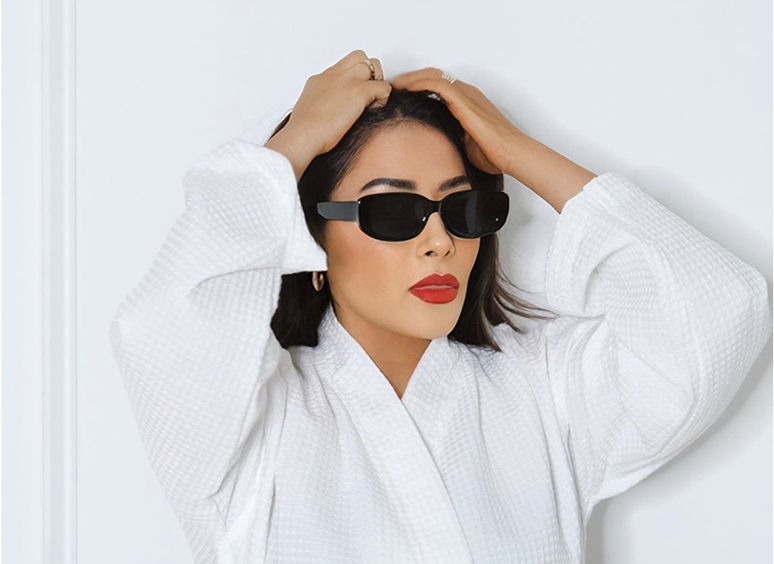 Woman wearing a white robe and black sunglasses, standing in front of a white wall
