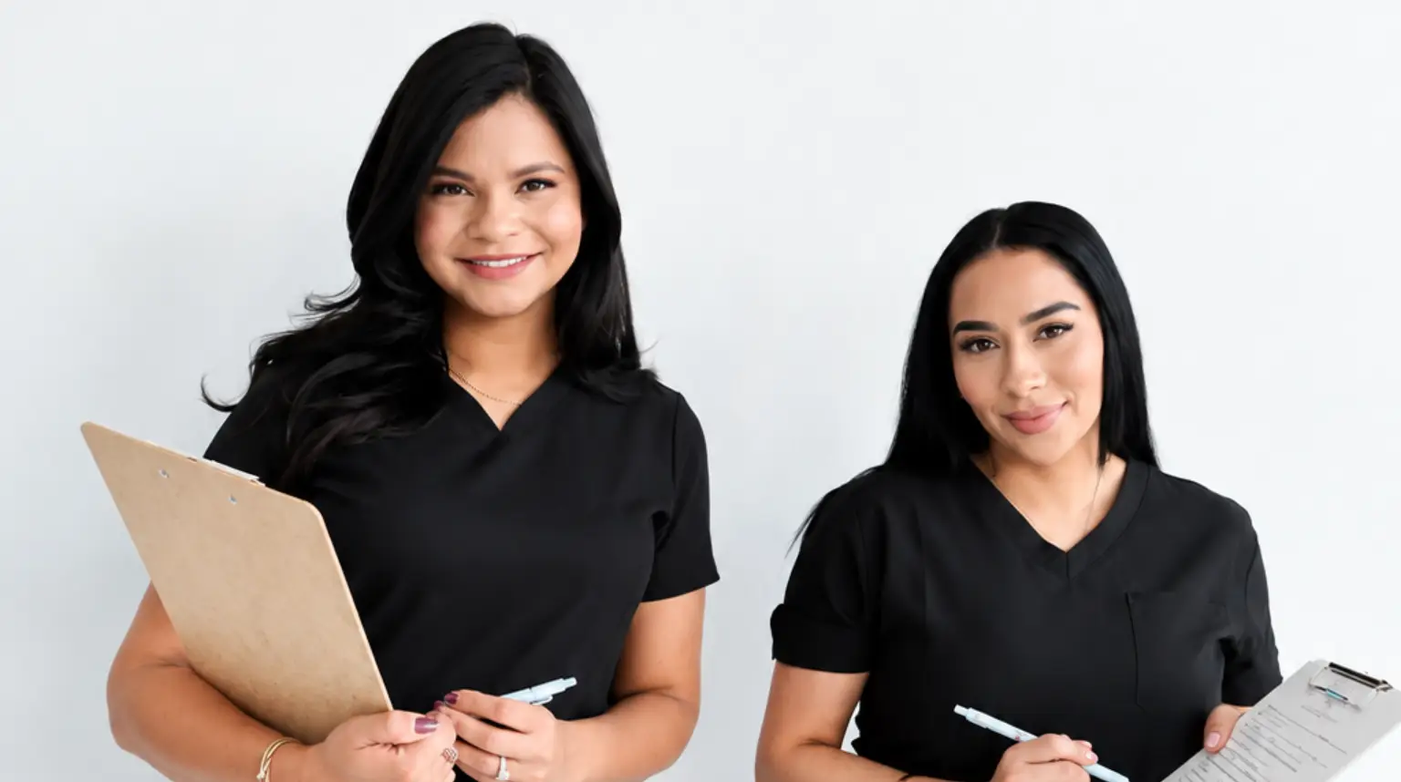 Two women in scrubs standing next to each other, each holding a clipboard and pen